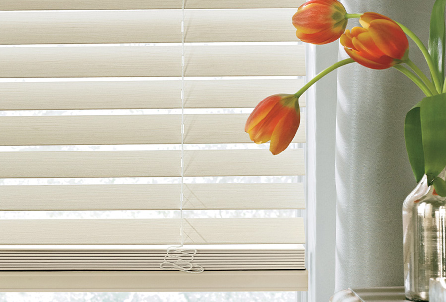 A vase of tulips sitting on a window sill next to faux wood blinds.